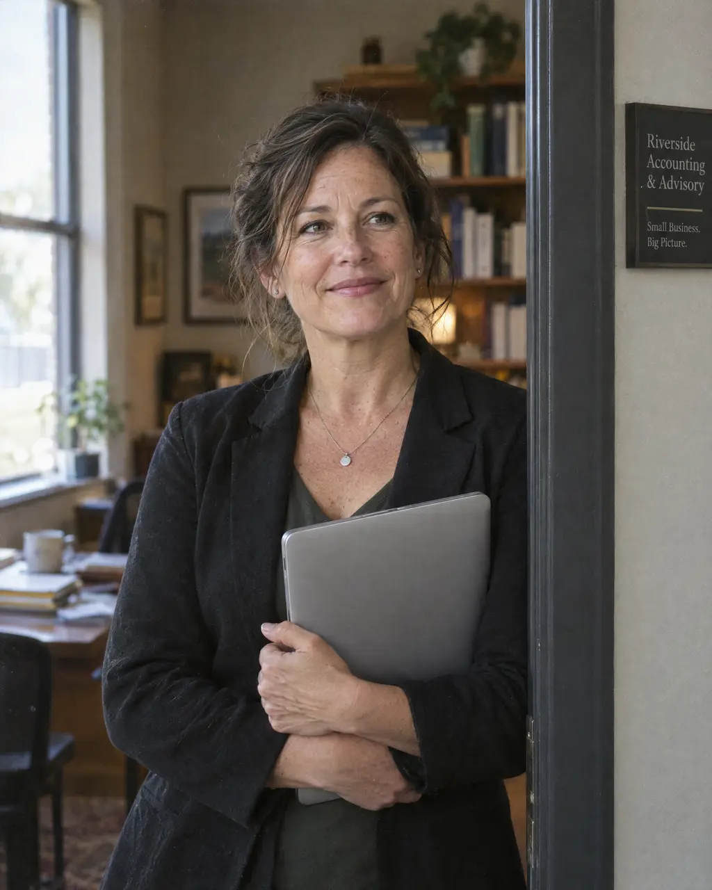 Owner of a small accounting firm standing in her office doorway holding a laptop